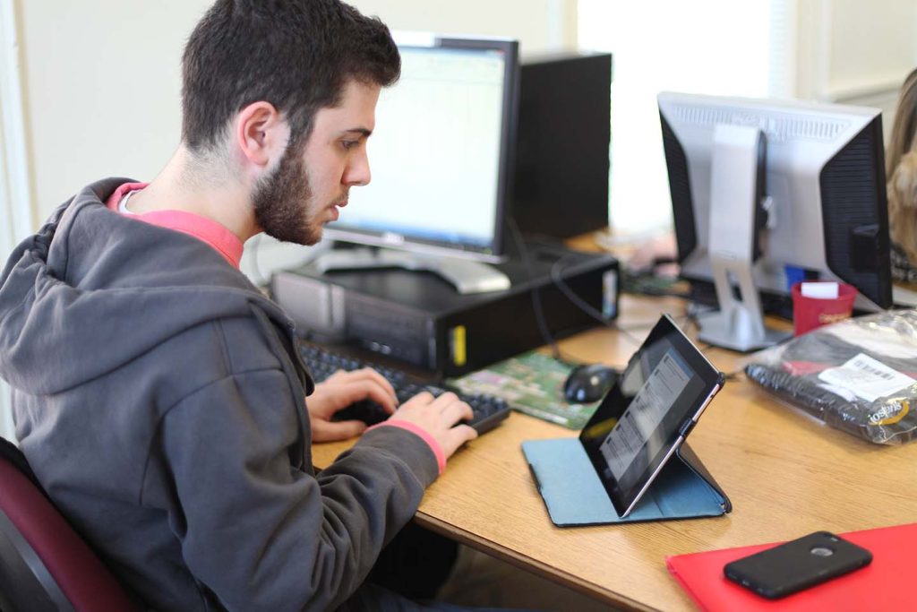 Student typing on keyboard while looking at tablet and a phone on the desk