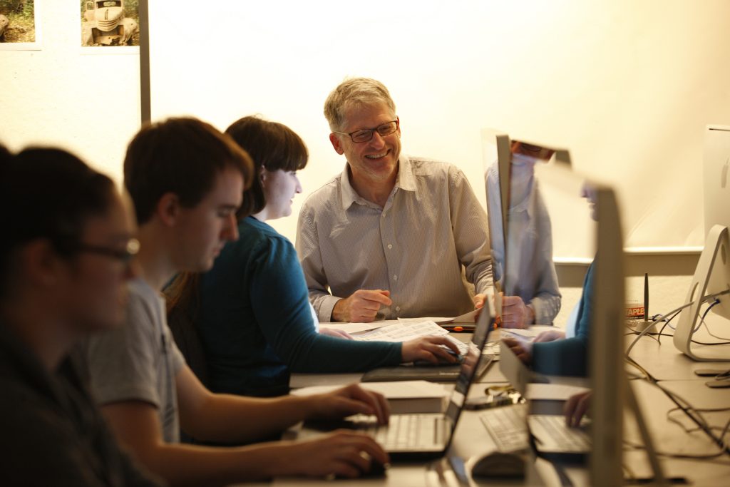 Professor looking at computer with class with students and computers