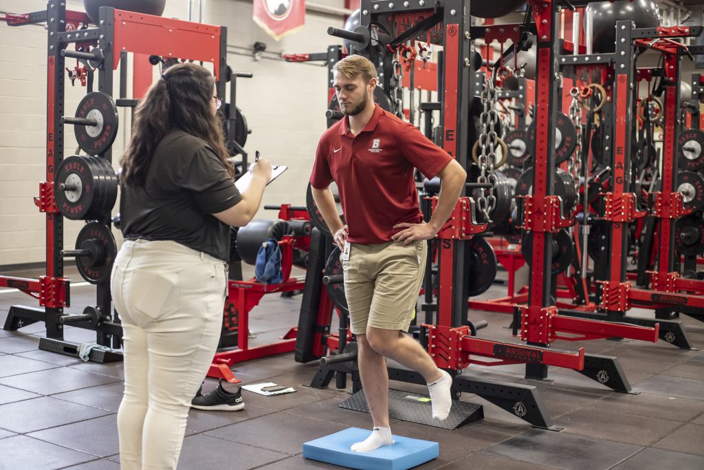 Student athletic trainer writes on clipboard while watching another student balance on one foot.