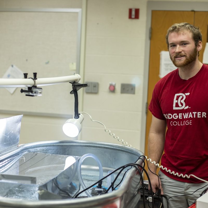 A male student stands by a fish tank he made to test water flow and current preference of longnose dace, a small freshwater fish. He is smiling and wearing a Bridgewater College t-shirt.