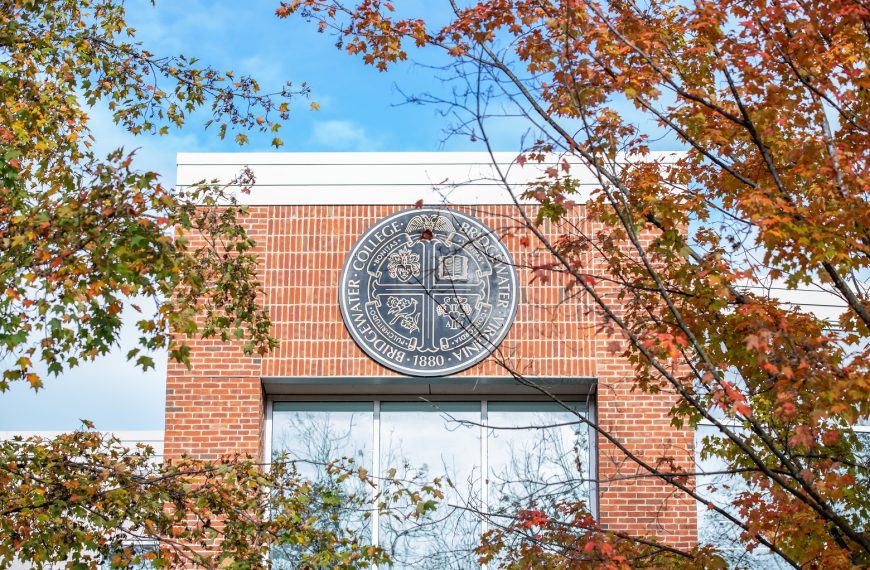 The Bridgewater College seal pictured on a brick building with fall tree leaves surrounding it