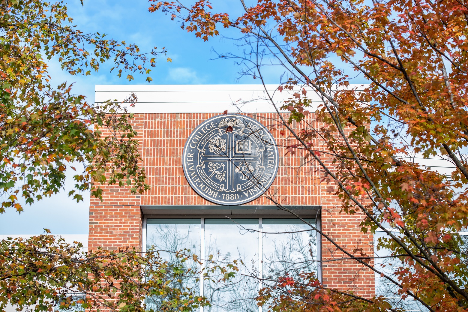 The Bridgewater College seal pictured on a brick building with fall tree leaves surrounding it