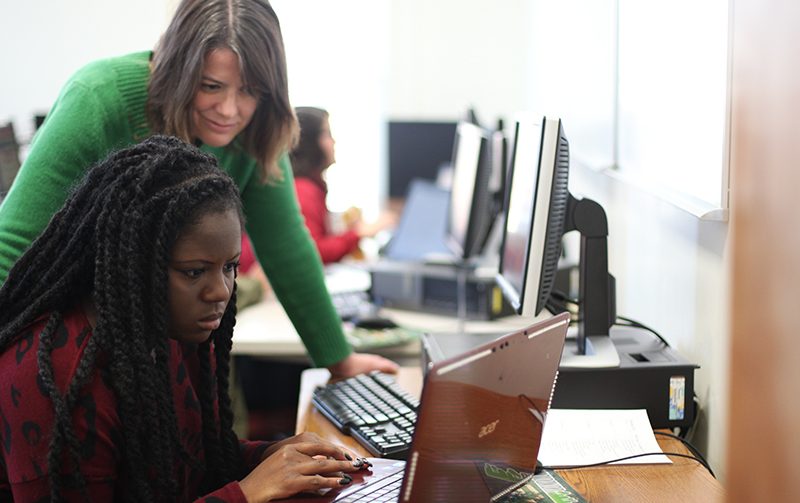 Communications professor standing behind student looking at computer