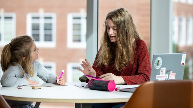 Student in teacher education program sitting at a desk working with a 2nd grader