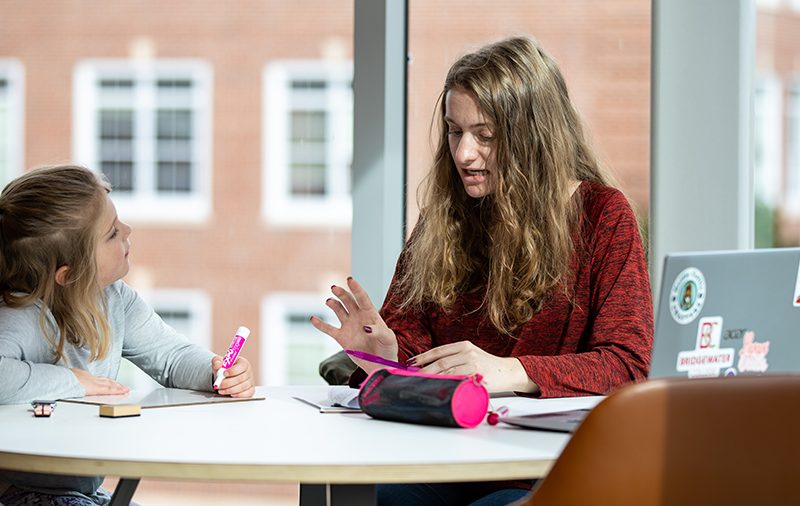 Student in teacher education program sitting at a desk working with a 2nd grader