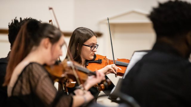 Student playing violin during performance surrounded by other players.