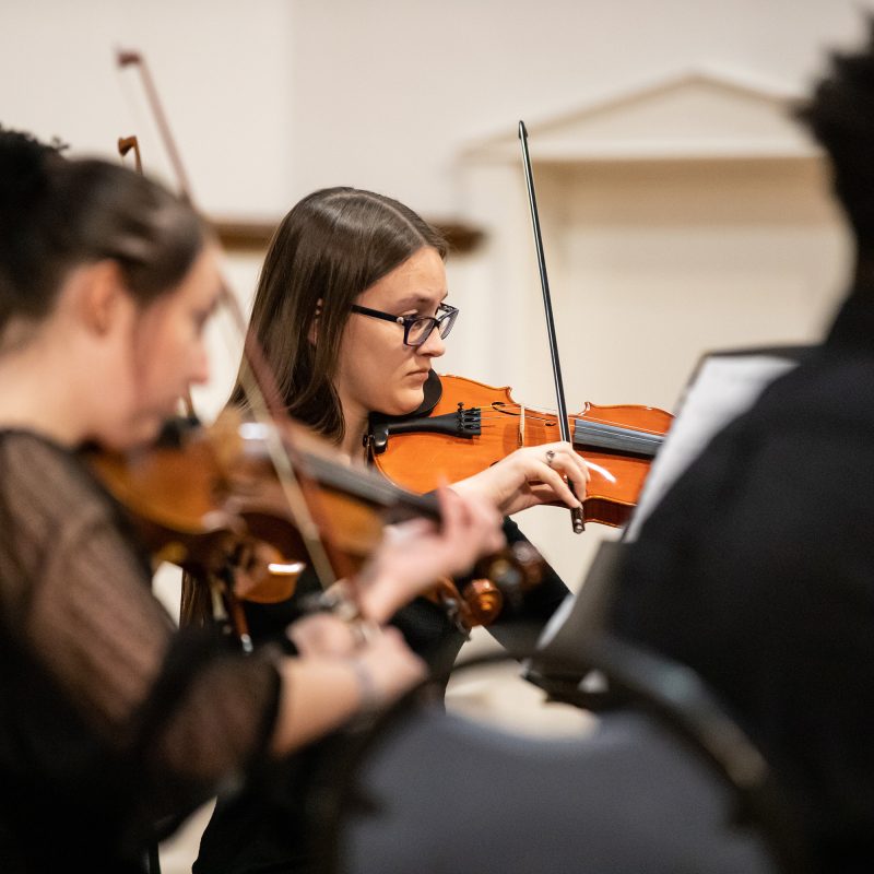 Student playing violin during performance surrounded by other players.