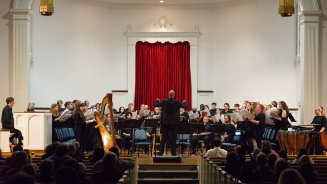 A music conductor stands at center stage with his arm raised and back to the audience. Members of the choir stand before him, facing the audience. The audience is seated in pews.