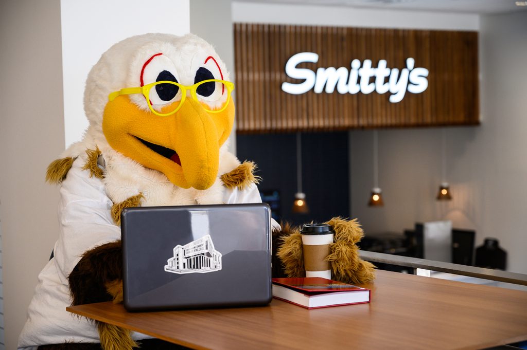 Mascot Ernie sitting with a laptop, coffee and a book with Smitty's in the background