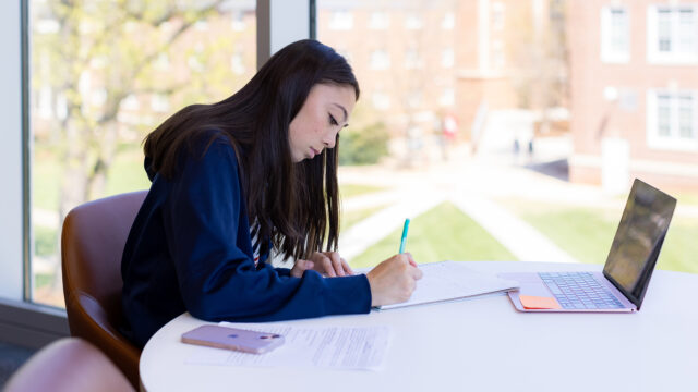 Student sitting at a desk writing on a piece of paper with a phone and laptop on the table.