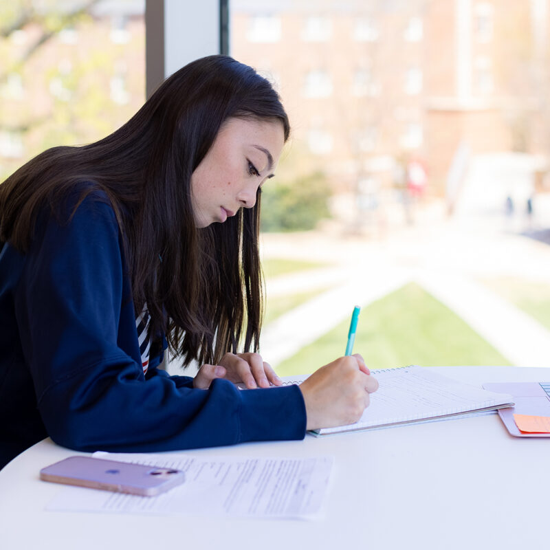 Student sitting at a desk writing on a piece of paper with a phone and laptop on the table.