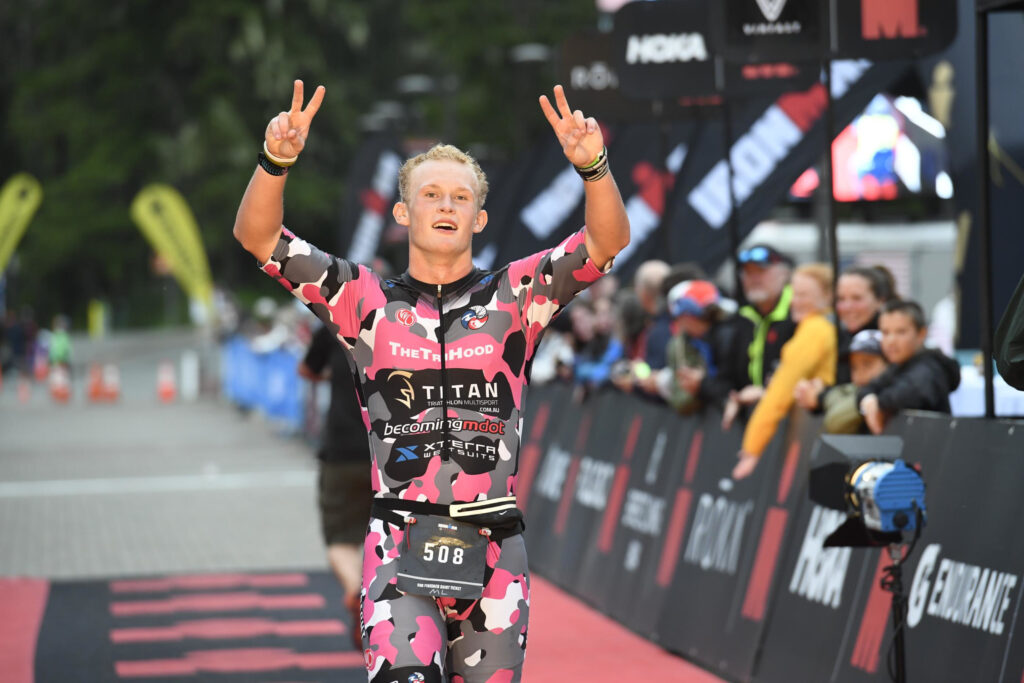 Grant Heidebrecht crossing the finish line of Ironman Alaska smiling and holding two peace signs in the air