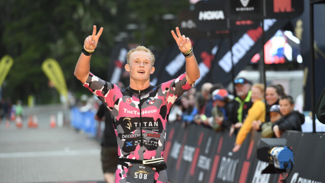 Grant Heidebrecht crossing the finish line of Ironman Alaska smiling and holding two peace signs in the air