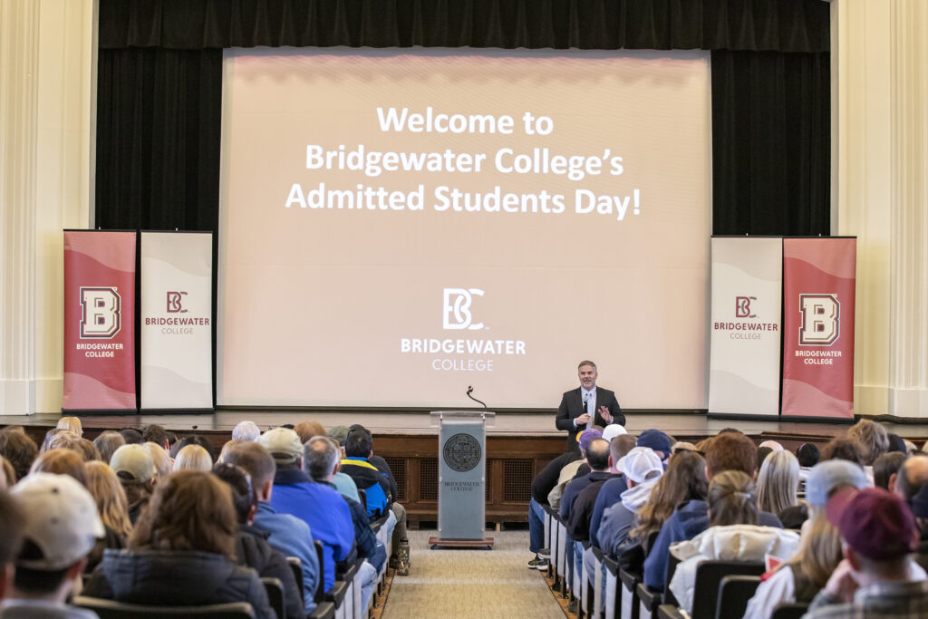 Vice President for Enrollment Management, Michael Post, standing in front of crowded Cole Hall with a projected image behind him that reads "Welcome to Bridgewater College Admitted Students Day"