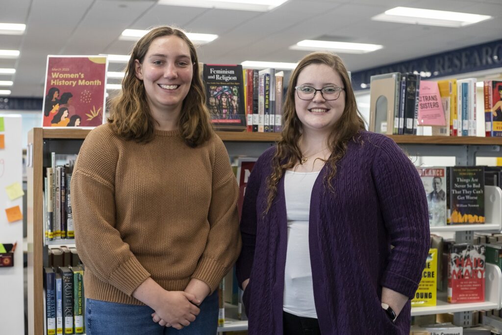 Two young women stand in front of a bookshelf in the library smiling