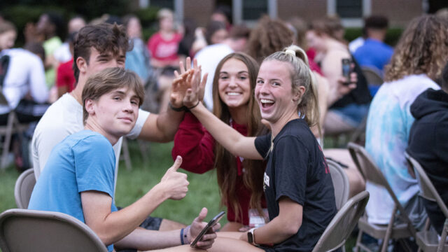 Group of boy and girls sitting in folding chairs forming a circle and high fiving in the middle. One boy on the left is giving a thumbs up to the camera.