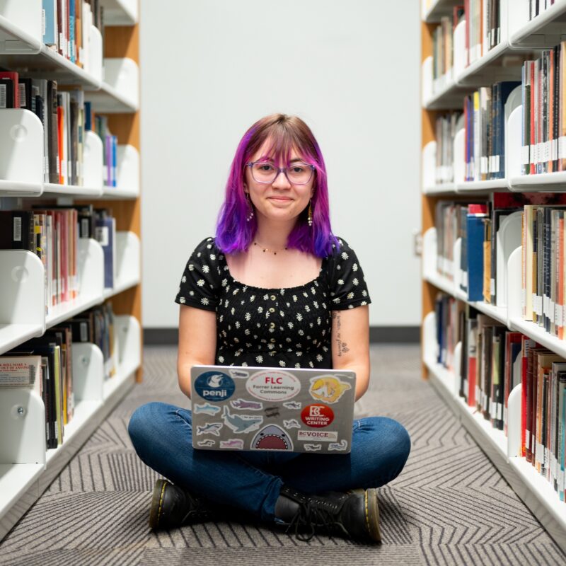 Mackenzie Hammack sitting on the floor between two bookcases with a laptop in her lap