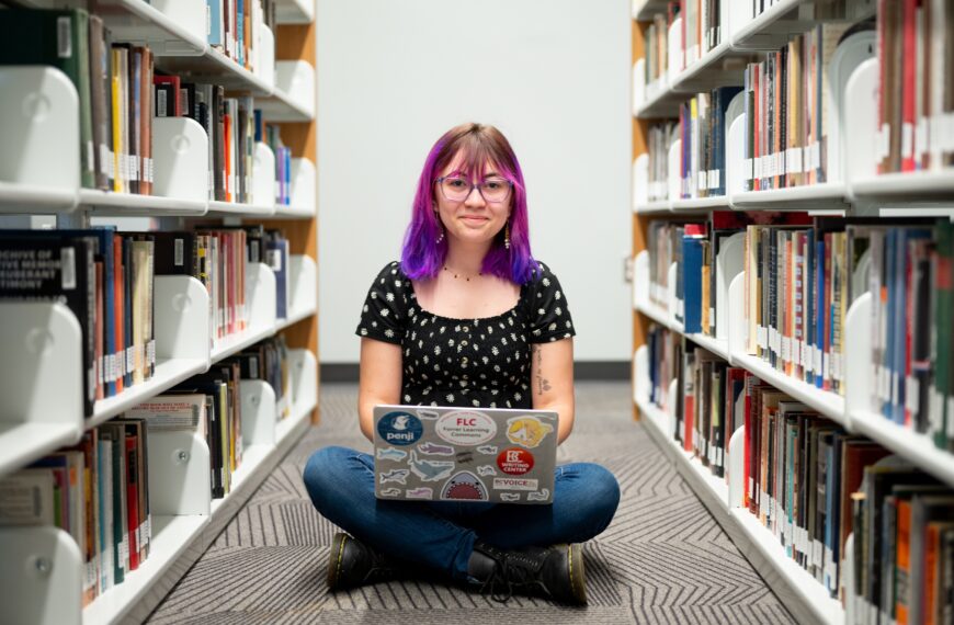 Mackenzie Hammack sitting on the floor between two bookcases with a laptop in her lap