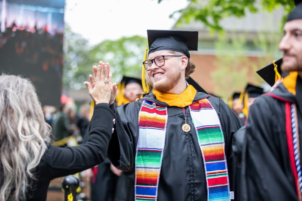 A graduate in regalia smiling and high-fiving someone in the crowd.