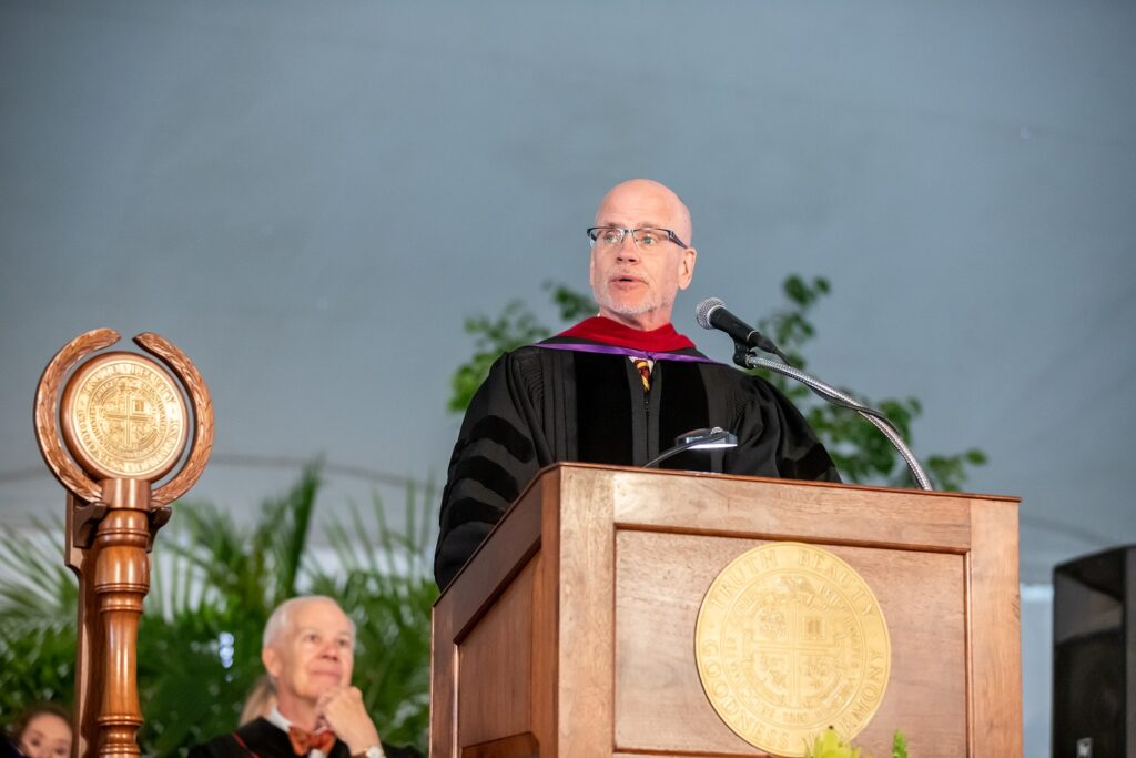 A man standing at a podium address the crowd. He is wearing academic regalis