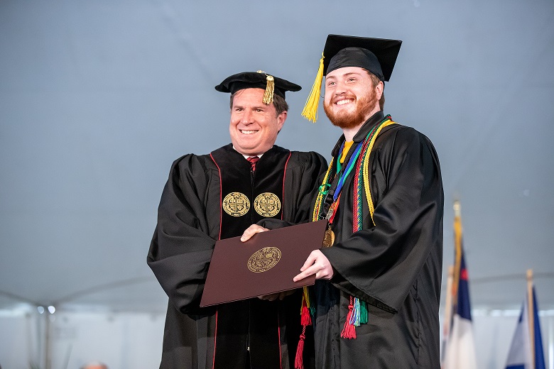 Two men in academic regalia smiling while receiving a diploma.
