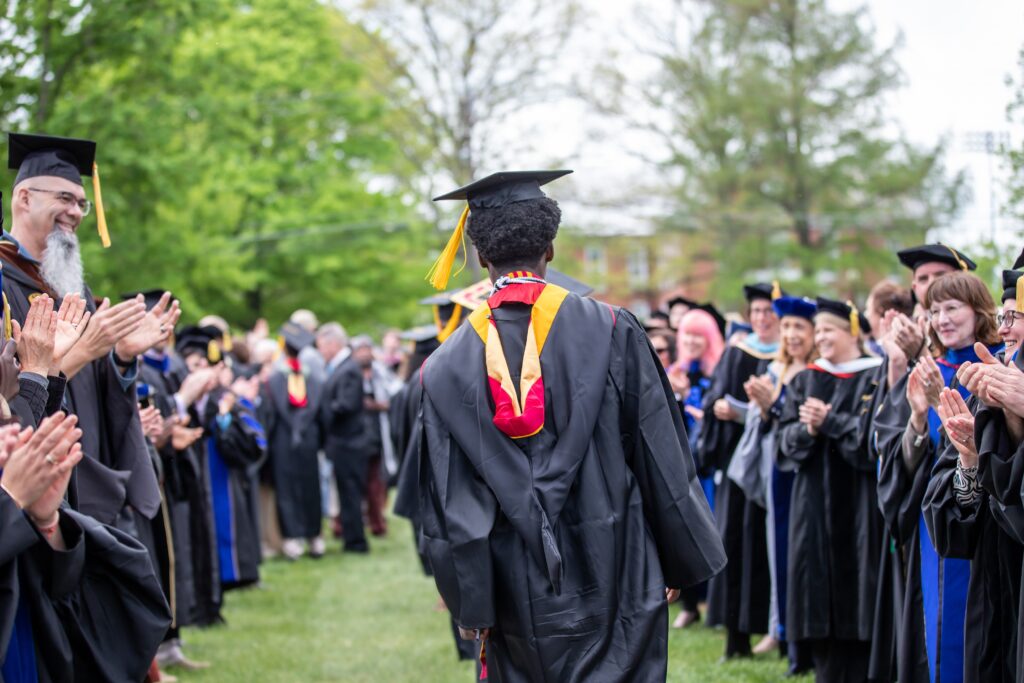 A student in a graduation cap and gown walks through an echelon of professors also in graduation regalia