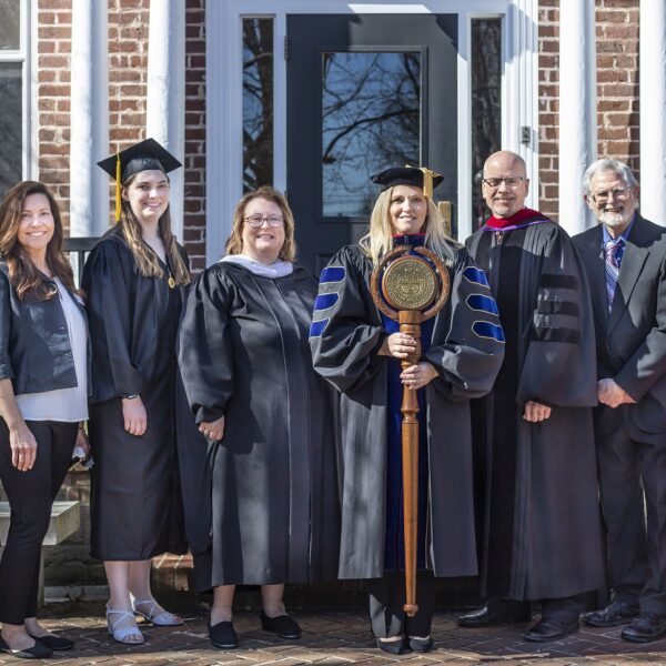 Eight men and women stand in front of a brick building smiling and wearing academic regalia.