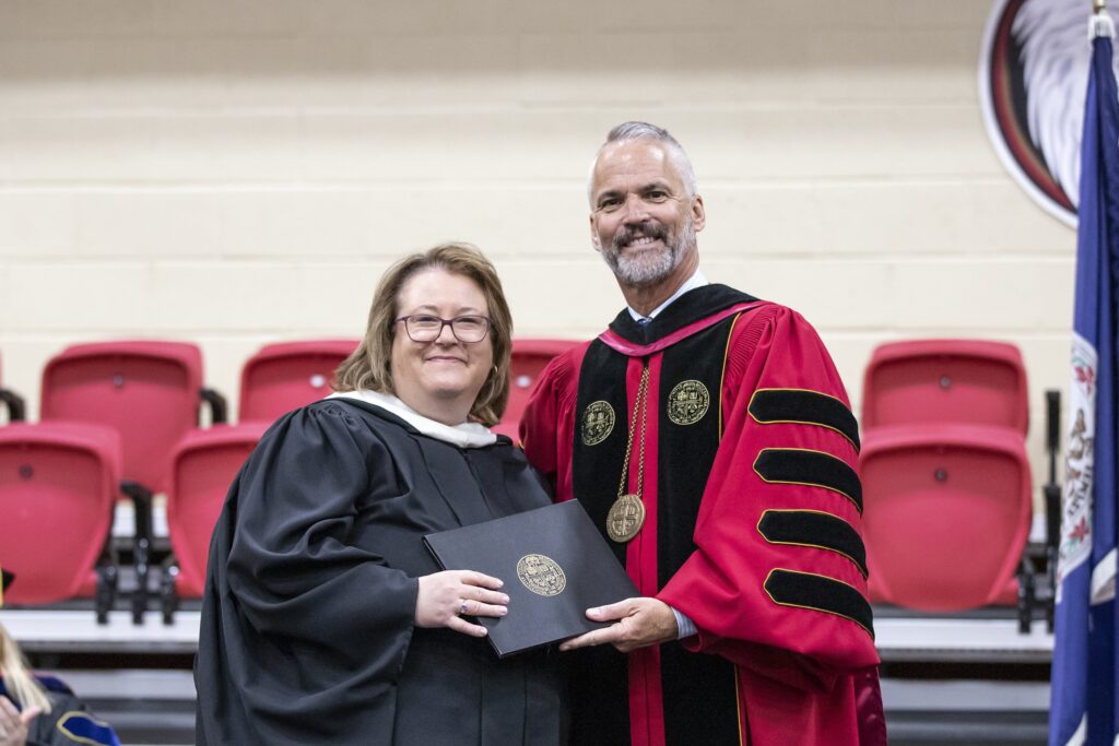 Jennifer Babcock and President David Bushman smiling together on stage in their academic regalia.