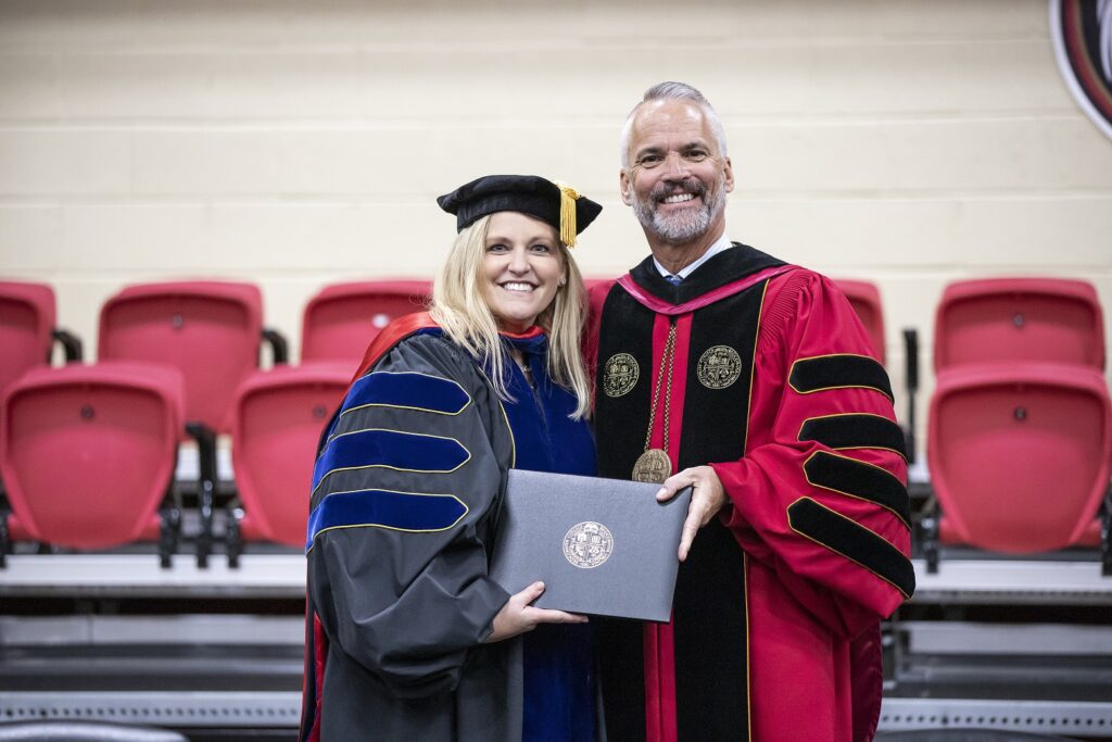 Donna Hoskins and President David Bushman smiling together on stage in their academic regalia.