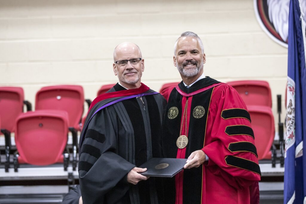 Robbie Miller and President David Bushman smiling together on stage in their academic regalia.