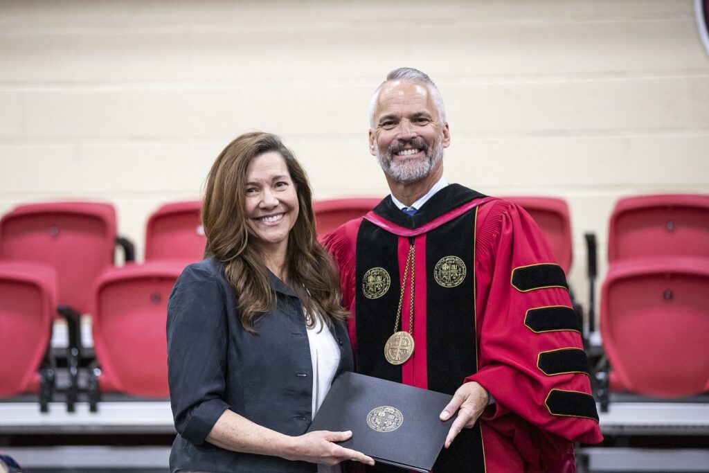 Shay Kelley and President David Bushman smiling together on stage in their academic regalia.