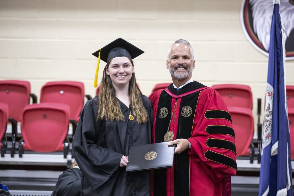 Hannah McPherson and President David Bushman smiling together on stage in their academic regalia.