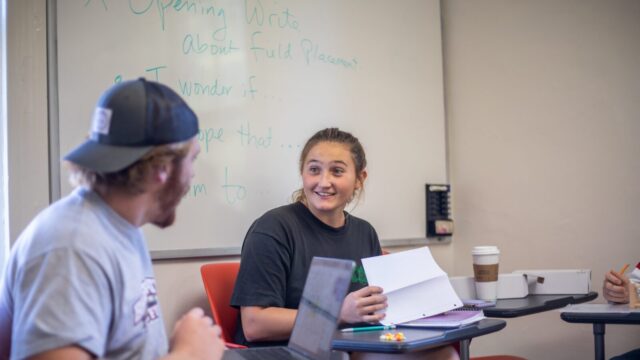 Student sitting at a desk with notebook in front of her as she turns and talks to a student in the foreground