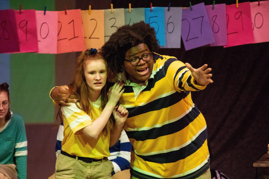 Two students in theatre production with one reaching out toward the audience and the other holding onto him looking concerned 