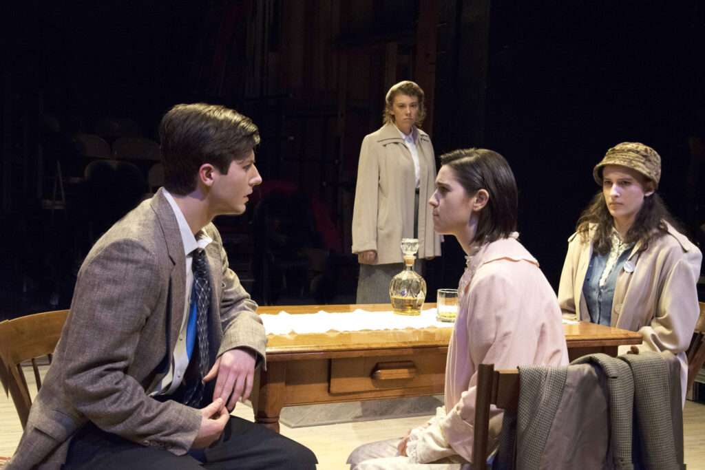 Four students performing in a theatrical production sitting around a table