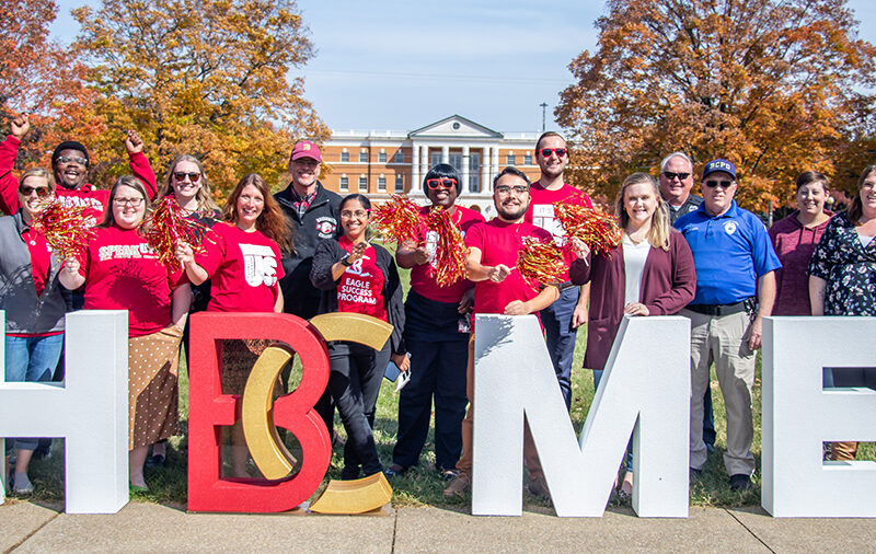 Large group of employees pose for a photo in front of letters that spell home with B-C to replace the O