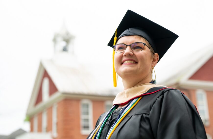 A female student in a cap and gown looking off into the distance while smiling. A brick building is in the background.