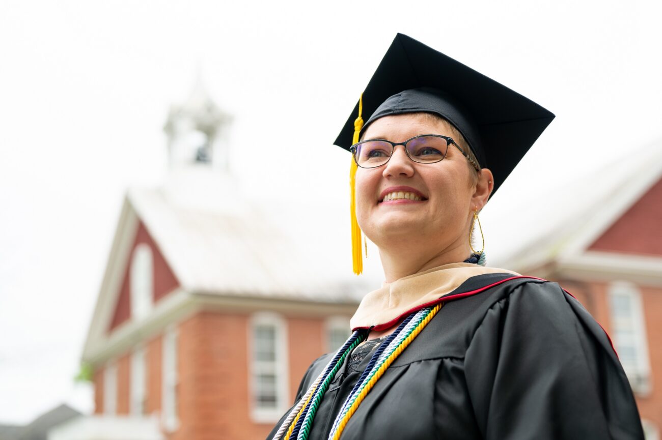 A female student in a cap and gown looking off into the distance while smiling. A brick building is in the background.