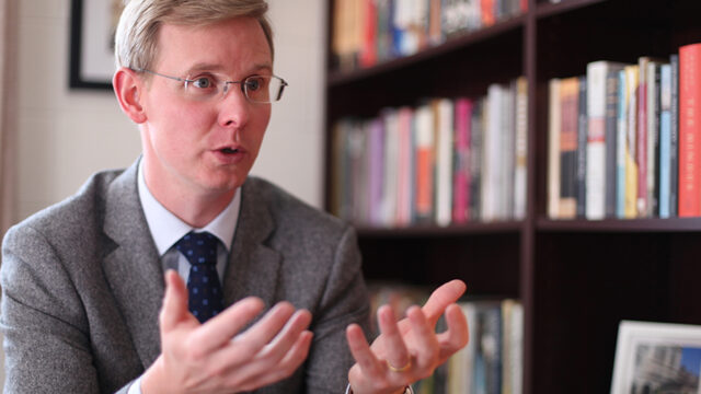 Professor speaking with hands out and a large bookshelf in the background