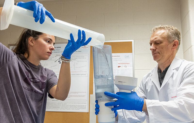 Student pouring a liquid into a tube a professor is holding for a summer research project