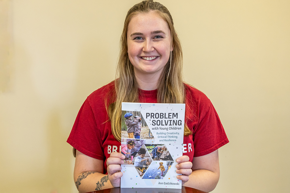 Student holding up a book titled "Problem Solving with Young Children" smiling for the camera