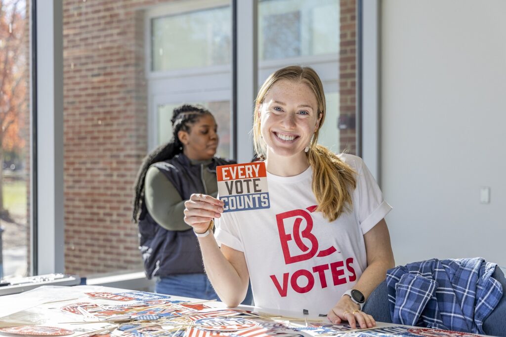 A female student wearing a B-C votes shirt and holding a sticker that says every vote counts