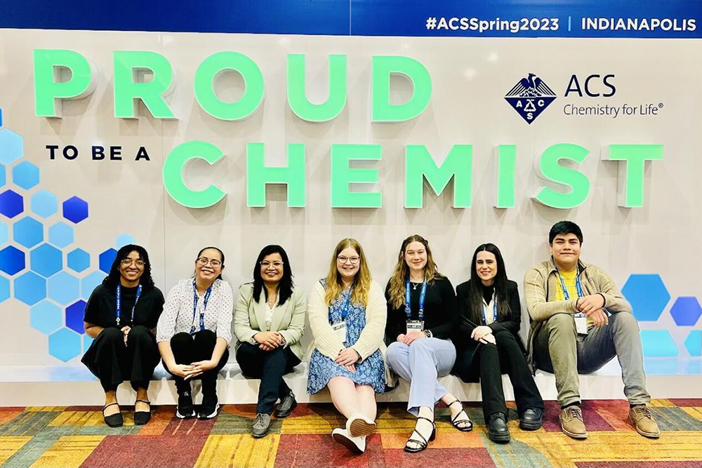 7 people sitting in front of large sign that says proud to be a chemist 