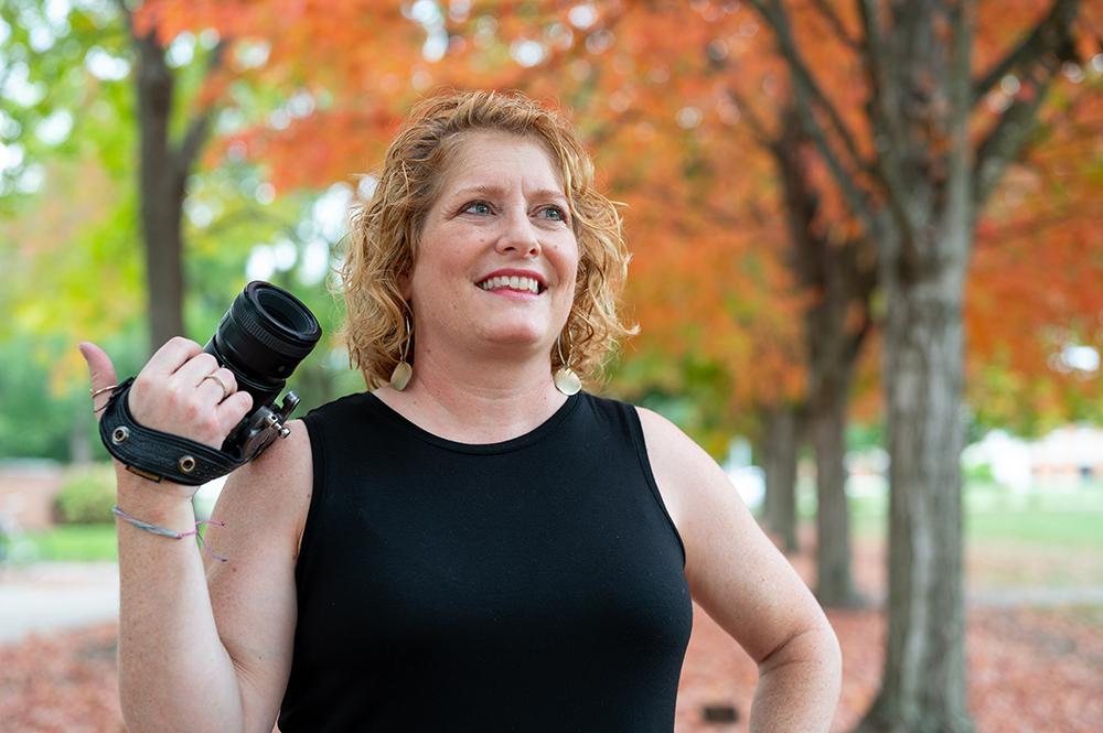 Woman stands holding a camera looking off in the distance with colorful fall trees in the background