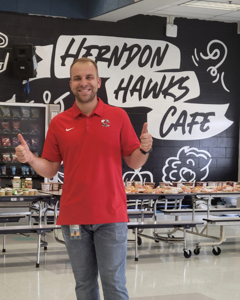 Gabe Segal posing for a photo giving two thumbs up in front of wall that says Herndon Hanks Cafe