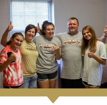 Family of five standing in a residence hall posing for a picture giving thumbs up