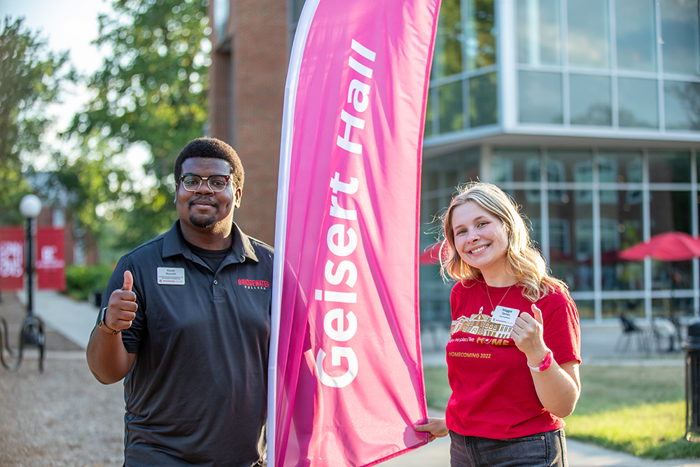 Resident Life giving thumbs up in front of flag that reads Geisert Hall