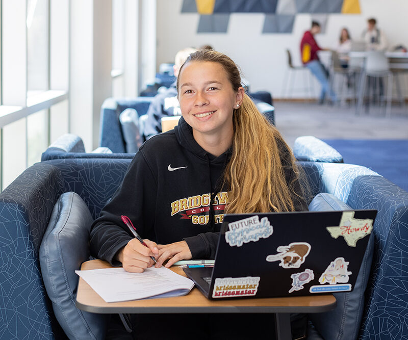 Student smiling for camera sitting at desk with paper and laptop