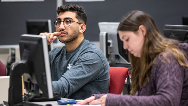 One student looking forward in class with another student in the foreground looking at the desk