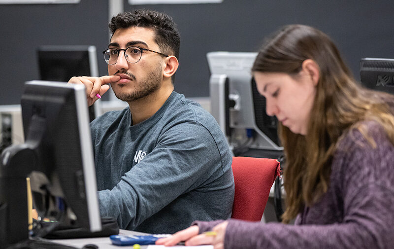 One student looking forward in class with another student in the foreground looking at the desk